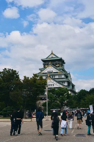青空と雲を背に立つ大阪城の天守と、そこへ向かう人たち